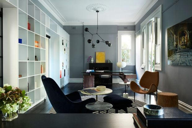 A refined living room or sitting room with grey walls, white built-in open shelving, dark timber flooring, and a compact mid-century-inspired furniture arrangement. The space includes sculptural lounge chairs, a dark coffee table, a desk or console, a television, striped rug, decorative artwork, and a contemporary pendant light. Tall windows bring in natural light, while ornate ceiling detailing and traditional cornices add architectural character.