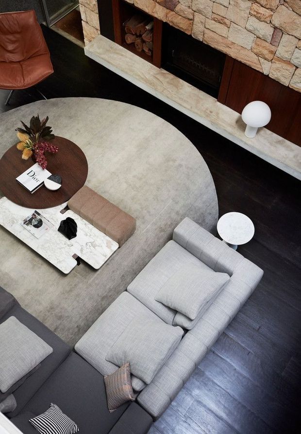 Overhead view of a refined living room centred on a large pale oval rug, grey sectional seating, a brown leather lounge chair, round timber coffee table, and a white marble coffee table. A substantial stacked-stone fireplace with dark timber cabinetry and a pale stone hearth forms the backdrop, complemented by dark timber flooring and minimalist table lamps.