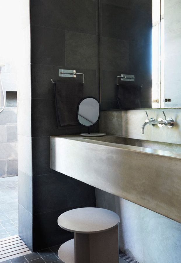 A contemporary bathroom featuring a long floating concrete vanity with wall-mounted chrome taps, dark large-format tiled walls, a broad mirror, black textured towel, and a small round stool. A partial shower area with pale tile is visible at left.