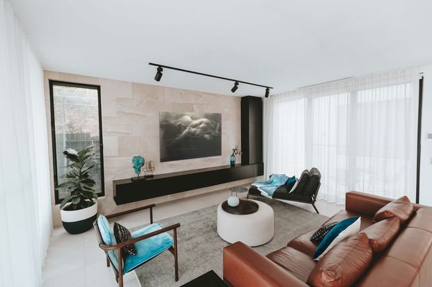 Bright contemporary living room with a large brown leather sofa, turquoise-accented lounge chair, round white coffee table, pale rug, sheer floor-to-ceiling curtains, and a black built-in media console. A pale stone-look feature wall frames the television, with black track lighting and expansive glazing creating an airy feel.