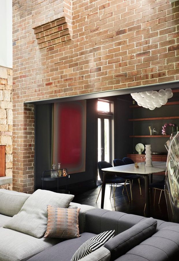 A stylish open-plan interior combining a living room and dining area. The space features exposed red brick walls, dark built-in shelving and cabinetry, a long dining table with navy upholstered chairs, and a large contemporary sectional sofa in the foreground. A sculptural clustered pendant light hangs above the dining table, while a bold red wall panel and decorative objects add contrast.