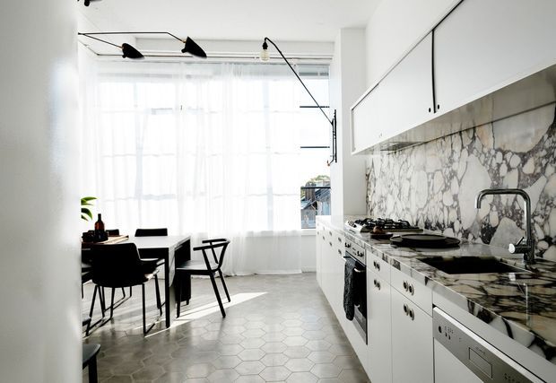 Bright open-plan kitchen and dining area with white cabinetry, a dramatic veined marble splashback and benchtop, pale hexagonal floor tiles, black accents, and full-height sheer curtained windows. A linear black track light and contemporary furnishings create a refined monochromatic interior.