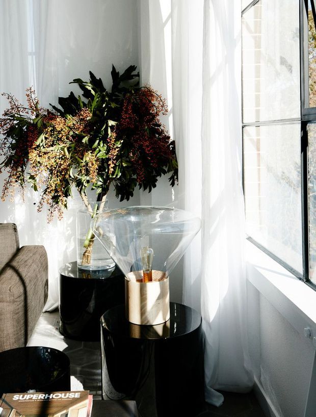A bright, refined interior corner beside a large black-framed window, with sheer white curtains, a grey upholstered armchair, glossy black cylindrical side tables, a clear glass vase containing leafy branches and berries, and a sculptural translucent glass lamp with a metallic cylindrical base. Strong natural light creates a calm, contemporary styled vignette.