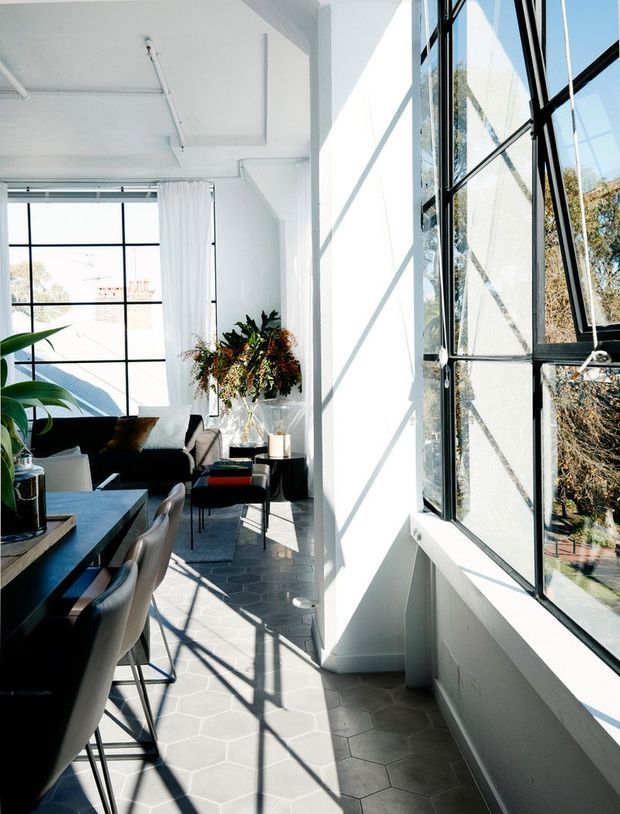 Bright, narrow contemporary living and dining space with expansive black-framed industrial-style glazing, white walls and ceiling, patterned grey hexagonal floor tiles, and strong geometric sunlight shadows. A dark dining table with upholstered chairs occupies the foreground, while a black sofa, coffee table, plants, and pendant-free minimalist furnishings sit beyond.