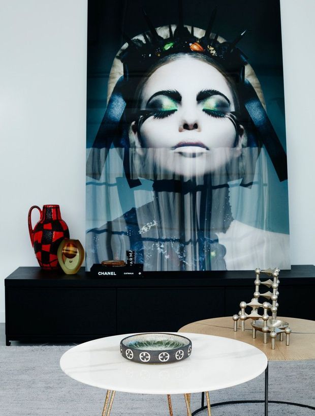 A stylish contemporary living room featuring a large dramatic fashion portrait artwork above a low black media console. The room includes a round white coffee table with a decorative bowl, a secondary round timber table, sculptural metallic candleholders, and bold decorative ceramics. The palette is predominantly black, white, grey, and muted timber, creating a refined, gallery-like atmosphere.