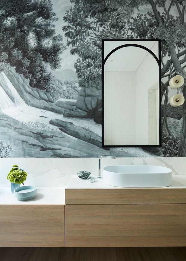 A refined contemporary bathroom vanity with a wall-mounted natural timber cabinet, white stone benchtop, and white oval vessel basin. A large black-framed mirror is set against a dramatic monochrome botanical and waterfall mural, with sculptural wall lights flanking it. The scheme combines warm wood, white surfaces, and dark botanical tones.
