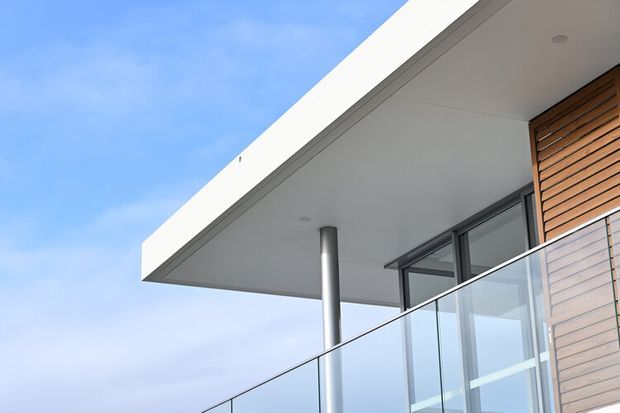 Close-up exterior architectural view of a contemporary upper-level balcony or terrace, featuring a broad white flat roof overhang, slim cylindrical metal support column, frameless glass balustrade, dark-framed glazing, and vertical timber wall cladding. The composition emphasizes clean horizontal and linear forms against a blue sky.