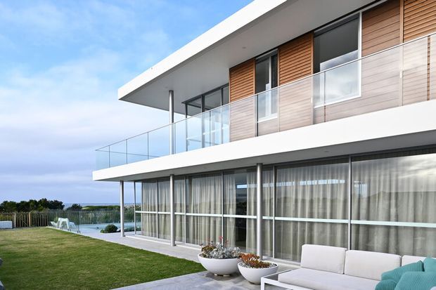 A contemporary multi-level residential building viewed from the exterior, featuring broad white horizontal slab edges, extensive floor-to-ceiling glazing, frameless glass balcony balustrades, and warm horizontal timber wall cladding. A covered ground-level terrace with outdoor seating opens onto a lawn, with a coastal-looking horizon visible beyond.