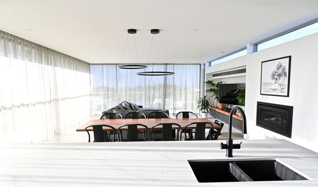 Bright open-plan kitchen and dining area with a large white stone island in the foreground, black undermount sink and tap, timber dining table with black chairs, a modern living area beyond, floor-to-ceiling sheer curtains, linear glazing, built-in shelving, and a contemporary fireplace.