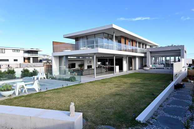 Exterior view of a large contemporary two-storey residence with a flat roof, extensive floor-to-ceiling glazing, projecting upper-level volumes, glass balustrades, and an open-plan ground floor connected to a landscaped lawn and pool terrace. The facade combines white rendered surfaces, dark window framing, and timber-toned screening.