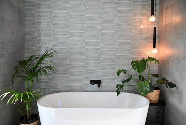 A contemporary bathroom featuring a freestanding white bathtub against a wall of pale grey elongated hexagonal tiles. Concrete-look grey wall finishes flank the tiled feature wall, with matte black tapware, warm pendant lights, and lush potted tropical plants adding contrast and softness.