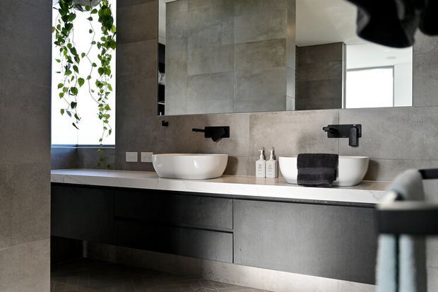 Contemporary bathroom vanity with two white vessel basins, matte black wall-mounted tapware, dark grey floating cabinetry, a pale stone benchtop, and large grey concrete-look wall tiles. A wide mirror reflects the room, while hanging greenery adds a natural accent.