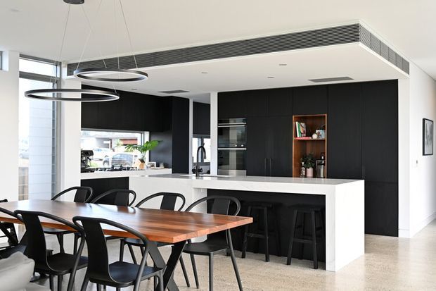 Open-plan contemporary kitchen and dining area with a large white island and breakfast bar, matte black full-height cabinetry, integrated appliances, timber dining table, black dining chairs and sculptural ring pendant lights. The space is bright and refined, with large windows and light flooring.