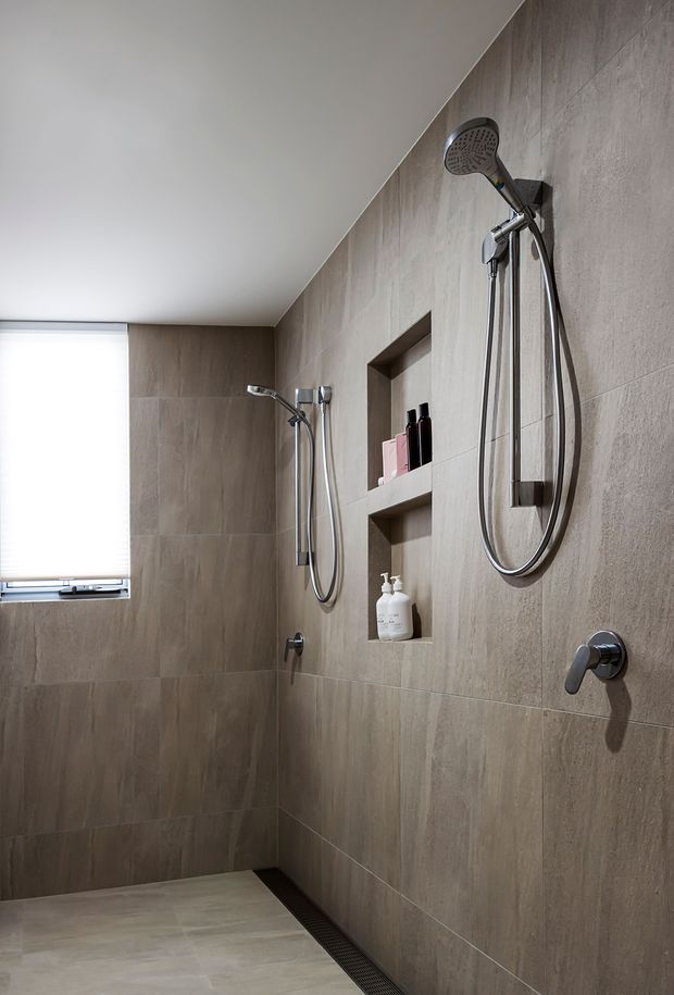 Contemporary walk-in shower with large-format taupe stone-look wall and floor tiles, recessed storage niches, chrome shower fixtures, concealed linear drain, and a small window with blind. The space has a clean, warm-neutral and minimalist appearance.