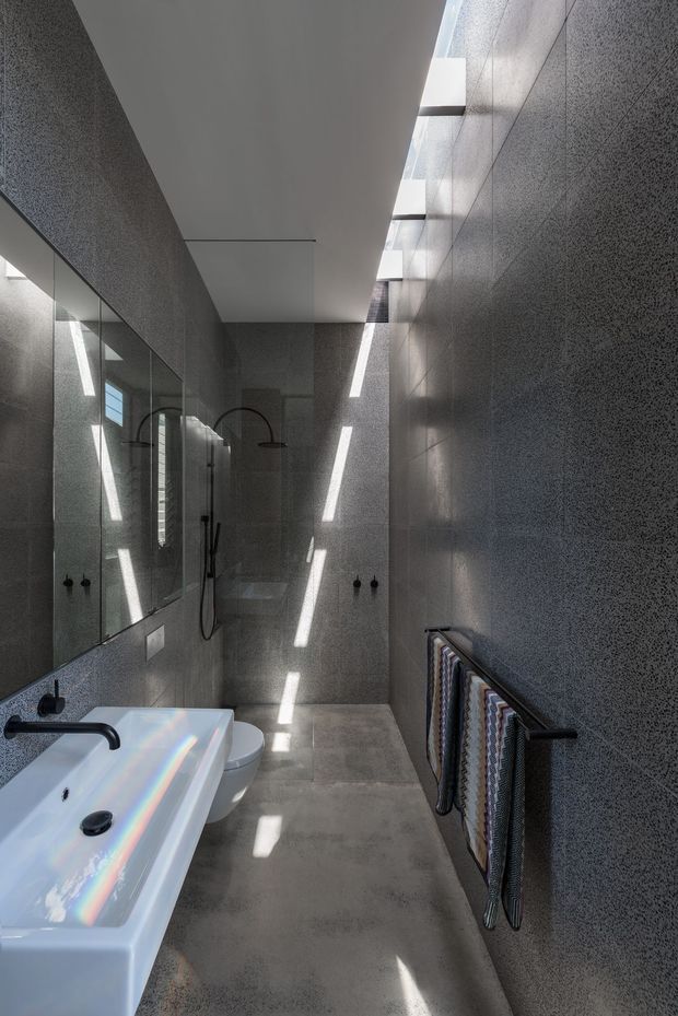 A narrow contemporary bathroom with dark grey textured wall surfaces, a large mirrored wall, concrete-look floor, white basin and toilet, and matte black tapware and shower fittings. Linear skylights or clerestory openings cast strong strips of daylight across the room. A heated towel rail with patterned towels is mounted on the right wall.