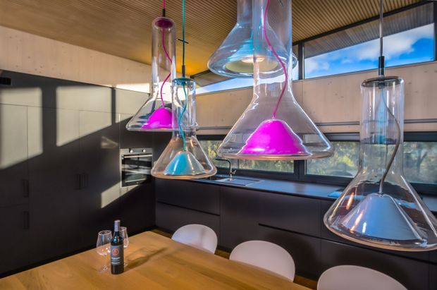 A contemporary kitchen and dining area with dark built-in cabinetry, integrated appliances, a warm timber dining table, and sculptural transparent glass pendant lights containing vivid magenta and blue accents. Floor-to-ceiling-style horizontal glazing brings in daylight, while a slatted timber ceiling adds texture.