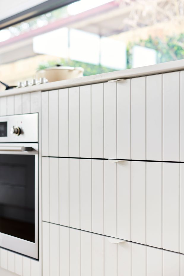Close-up view of a bright contemporary kitchen, focused on white vertically fluted cabinetry with integrated curved finger pulls. A built-in oven is visible at the left, with a pale benchtop and cooktop above. Large glazing and greenery appear softly in the background.