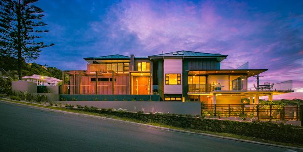 Twilight exterior photograph of a large multi-level contemporary residence on a sloping suburban site. The home features broad glazing, warm interior illumination, projecting roof planes, balconies and terraces, dark horizontal cladding, and substantial stone and rendered boundary walls. Landscaping and neighbouring structures are visible around the property.