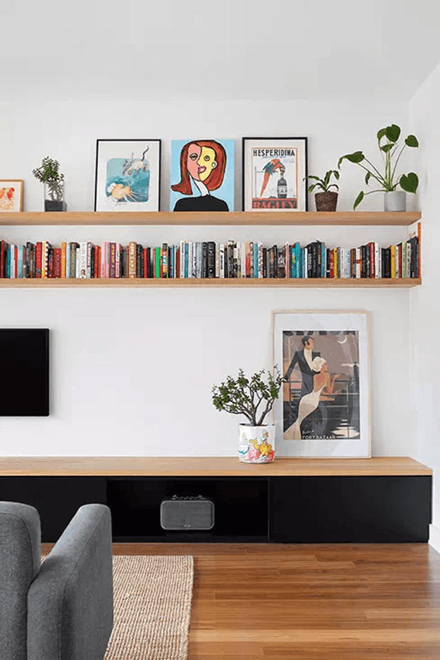 A bright, neatly styled living room media wall with two long timber shelves displaying books, framed artwork, and potted plants. A low black built-in entertainment cabinet with a timber top sits below, accompanied by a wall-mounted television, grey upholstered seating, and warm timber flooring with a woven rug.