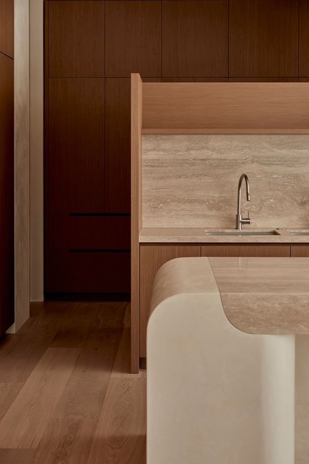 A close-up view of a refined, contemporary kitchen featuring a sculptural curved island with a pale stone benchtop and waterfall edge, integrated sink and brushed metal gooseneck tap. Warm timber cabinetry and matching timber flooring contrast with a full-height beige stone splashback and horizontal timber overhead detail. A dark timber-lined passage or joinery wall is visible beyond the kitchen.