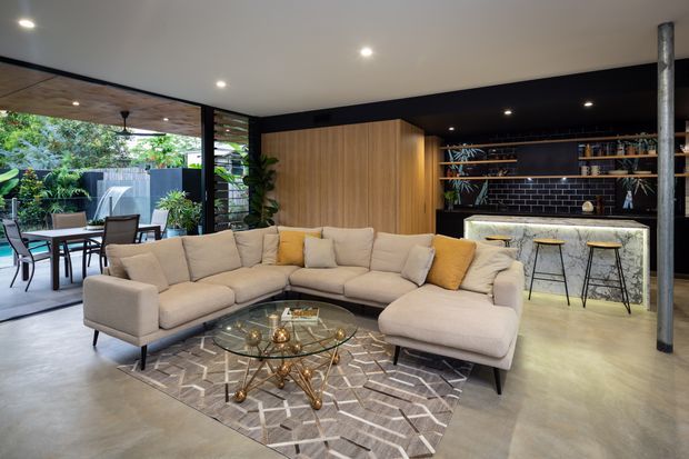 Open-plan contemporary living room with a large U-shaped cream sectional sofa, glass coffee table, patterned area rug, and integrated black bar or kitchenette. Full-height glazing opens to a covered outdoor dining area and lush garden. Warm timber joinery, black cabinetry and backsplash, marble-look bar front, recessed lighting, and a central structural post create a refined indoor-outdoor setting.
