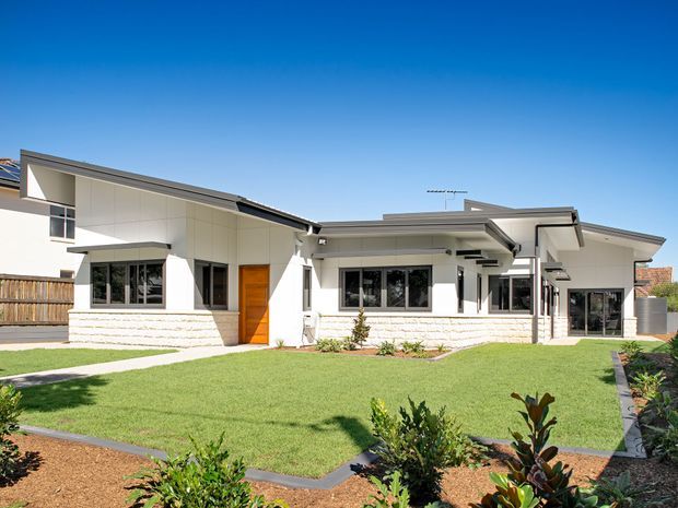 A bright contemporary single-storey residence viewed from the landscaped front yard. The home has a low-pitched, asymmetrical roofline, crisp white rendered walls, pale horizontal masonry at the base, extensive dark-framed glazing, timber entry doors, and projecting covered elements. A manicured lawn, garden beds, concrete paths, and timber fencing surround the facade under a clear blue sky.