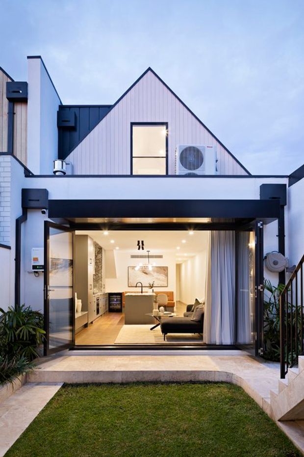 Exterior view of a compact multi-level contemporary home with a steep front gable clad in pale vertical weatherboard, dark metal accents, and a broad upper-level terrace or balcony. Large fully opened glazed doors reveal a bright open-plan interior with a kitchen island, dining area and living room. A small landscaped lawn and paved entry sit in the foreground, with stairs and planting framing the entrance.