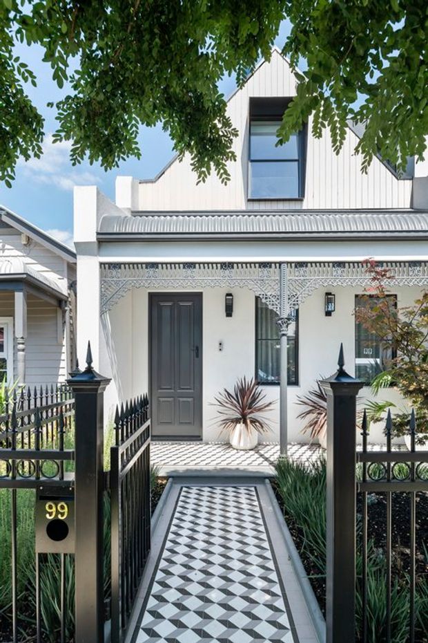 Street-facing view of a narrow heritage-style townhouse facade with a covered front porch, ornate white lacework and decorative columns, black metal fencing and gate, a patterned geometric tiled path, and a contemporary upper-level gabled extension clad in vertical white boards. Lush overhanging trees frame the facade.