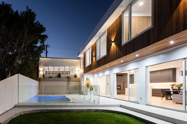 Dusk exterior view of a contemporary multi-level residence with a long white rendered facade, dark vertical timber cladding, extensive floor-to-ceiling glazing, recessed lighting, and a recessed swimming pool in a paved courtyard. Warmly lit interior living and dining areas are visible through the glazed doors, with landscaped lawn and boundary fencing in the foreground.