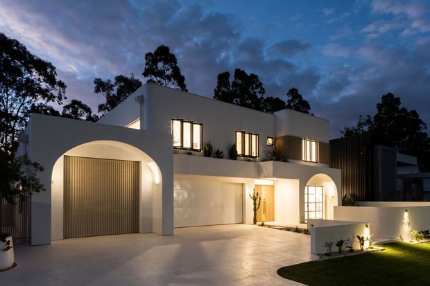 Twilight exterior photograph of a contemporary multi-level residence with smooth white rendered walls, rounded arched openings, a large arched garage entrance, tall illuminated windows, and a broad pale driveway. Dark vertical cladding appears on the right side, with landscaped lawn and architectural lighting in the foreground.