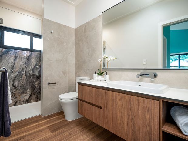Bright contemporary bathroom with a floating timber vanity, white integrated basin, large frameless mirror, wall-mounted tapware, tiled shower area, toilet, and warm wood-look flooring. A dramatic dark stone-look shower wall provides contrast, while a turquoise-painted doorway is visible in the mirror and at right.
