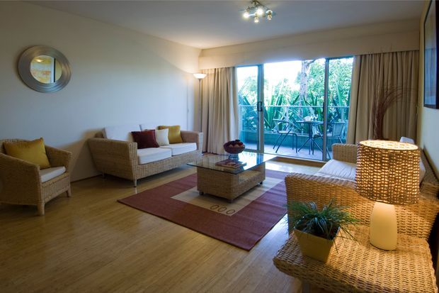 A warm, inviting living room furnished with woven rattan armchairs, a matching sofa, coffee table, and console table. The room has light timber flooring, neutral walls, full-height beige curtains, a patterned area rug, and a large woven table lamp. Sliding glass doors open to a leafy outdoor balcony or terrace, bringing in natural light.