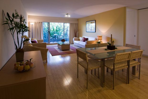 Open-plan living and dining area with warm timber flooring, woven rattan furniture, neutral upholstered seating, a glass-topped dining table, and a mustard-yellow feature wall. Large glazed doors open toward a leafy outdoor area, bringing natural light into the room. The space has a relaxed, warm, tropical-influenced interior character.