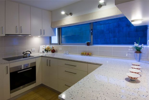 Bright, compact contemporary kitchen with pale flat-panel cabinetry, stainless-steel appliances, a speckled light benchtop, and a tiled splashback. A wide window brings in blue-toned exterior light, while a peninsula or island in the foreground provides additional preparation space and displays small bowls.