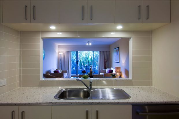 A compact galley-style kitchen viewed from the sink, with pale grey built-in overhead and base cabinetry, long dark handles, a speckled light benchtop, tiled splashback, and a stainless-steel double-bowl sink. A wide serving opening looks through to a furnished living room with large glazed doors or windows and curtains. Under-cabinet lighting illuminates the work area.