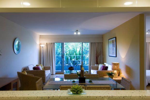 A warmly lit open-plan living and dining area viewed from behind a pale stone kitchen or serving counter. The room features woven rattan seating, a glass-topped dining table, a low coffee table, framed artwork, layered curtains, recessed and lamp lighting, and full-height glazed doors opening to a leafy outdoor setting. The interior has a relaxed resort-like feel with cream walls and natural woven textures.