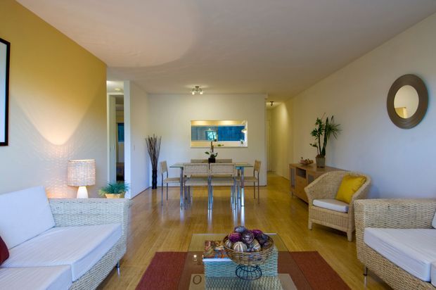 Bright open-plan living and dining area with white wicker seating, a glass-topped dining table, warm timber flooring, pale walls, and a yellow accent wall. A large circular wall mirror, indoor plants, woven furnishings, and a low coffee table create a relaxed contemporary coastal feel.