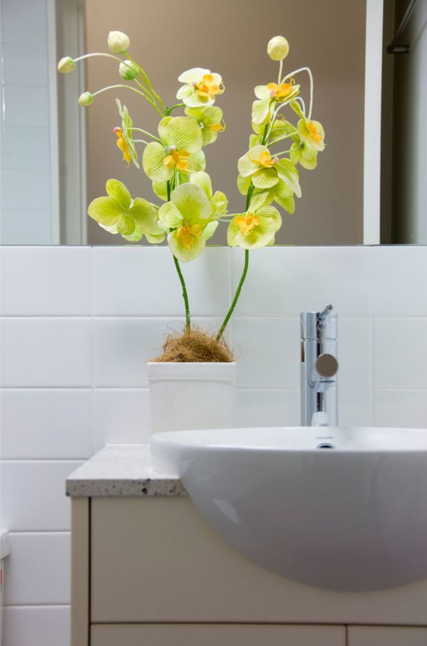 Close-up of a contemporary bathroom vanity featuring a white wall-mounted basin, chrome mixer tap, pale speckled benchtop, light cabinetry, and white rectangular wall tiles. A bright lime-green orchid arrangement in a white planter sits on the vanity beneath a large mirror.