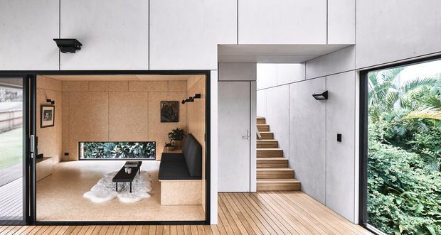 A minimalist modern interior showing a compact living room enclosed by dark-framed glazing, with a black sofa, low coffee table, pale rug, and warm wood-toned walls. In the foreground, a timber staircase leads upward beside smooth pale concrete or plaster wall panels. Large openings provide views to lush greenery outside, and linear wall-mounted lighting reinforces the restrained architectural design.