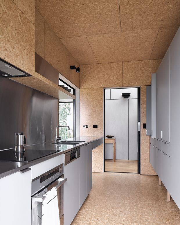 A compact contemporary galley kitchen with pale grey handleless cabinetry on both sides, a dark stainless-steel splashback and benchtop, integrated appliances, and a warm tan engineered-board finish covering the walls, ceiling, and floor. A glazed side opening brings in daylight, while a black-framed doorway leads to a secondary space with matching light timber flooring. The design is streamlined and minimalist with strong material continuity and dark hardware accents.