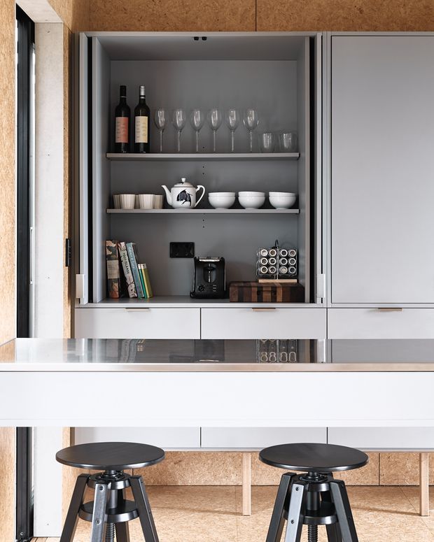 A compact contemporary kitchen with a built-in open display cupboard containing wine bottles, glassware, crockery, books and small appliances. White and grey cabinetry contrasts with warm wood-toned textured wall and panel surfaces. A reflective light benchtop extends into a peninsula or breakfast bar with two black stools. The space is neatly organised and softly lit, with a minimalist yet warm material palette.