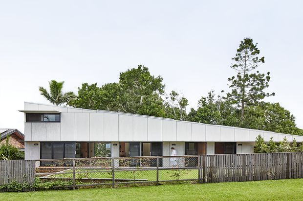 A long, low-slung contemporary residential building viewed from across a lawn. The facade uses pale grey-white vertical-panel cladding with dark-framed windows and glazed doors, and features a distinctive gently sloping roofline. A fenced front yard and mature tropical/subtropical vegetation surround the building.