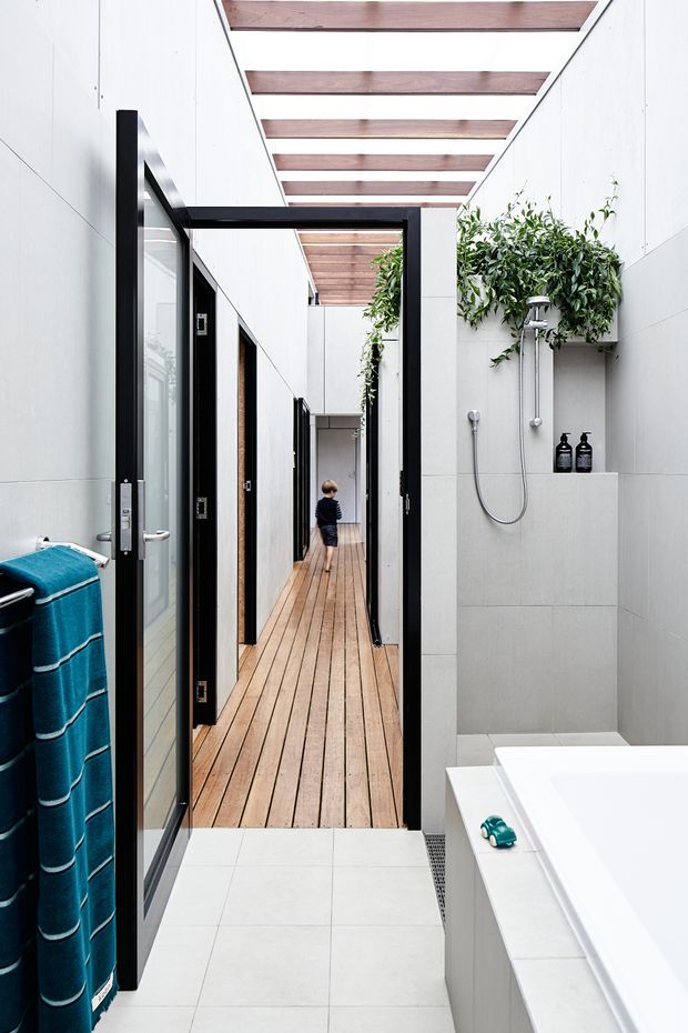 A narrow, bright contemporary bathroom opening onto a long timber-lined passage or outdoor shower court. The bathroom has large pale wall and floor tiles, a white built-in bathtub, a frameless glass shower enclosure with black metal framing, and a wall-mounted outdoor-style shower beneath a leafy planter. The passage continues toward another room and is covered by a slatted timber-and-white overhead canopy. A child is visible in the distance.