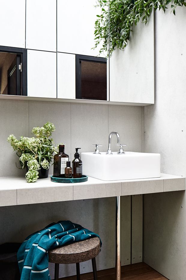 A compact contemporary bathroom vanity with a white vessel basin and polished chrome wall-mounted tapware. The vanity is clad in pale grey-beige tiled panels with a floating tiled benchtop, mirrored wall cabinets above, and an open lower section. A leafy potted plant, toiletries, and a small upholstered stool add warmth, while dark timber flooring and black-framed mirror cabinet details provide contrast.