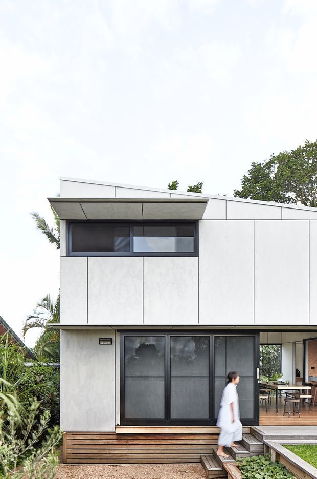 Exterior view of a compact contemporary two-storey house with a sharply angled roofline, white panelled facade, dark-framed windows and doors, and a timber deck and stepped entry landscape. The open ground-floor area reveals a dining space through the glazing, while a person walks down the timber steps.