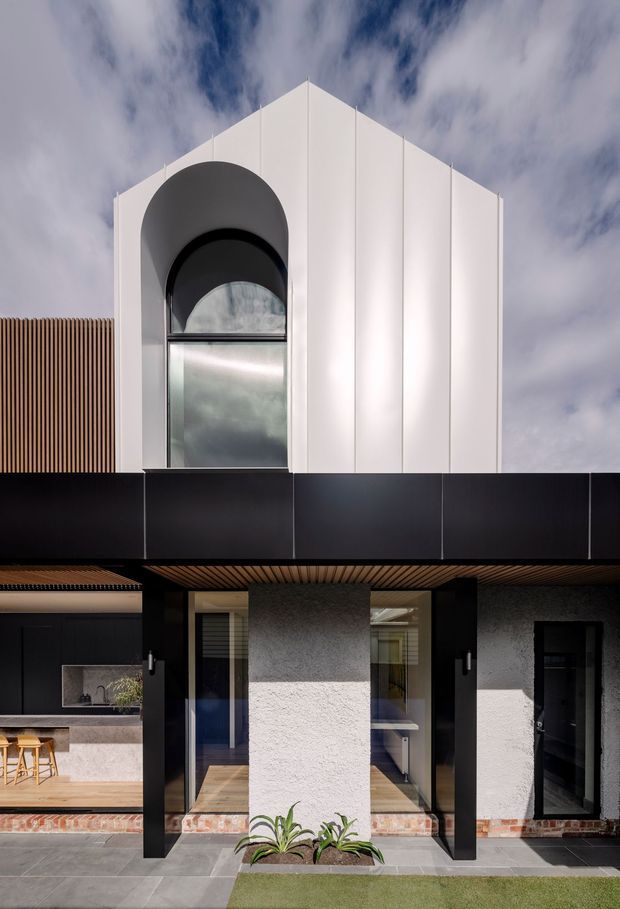 Front exterior view of a contemporary multi-level residence with a strong geometric gabled facade. The upper storey is clad in white vertical panels and features a tall arched window, while the lower level uses dark cladding, black structural columns, textured render, timber soffit screening, extensive glazing, and a paved entry terrace with minimal planting.