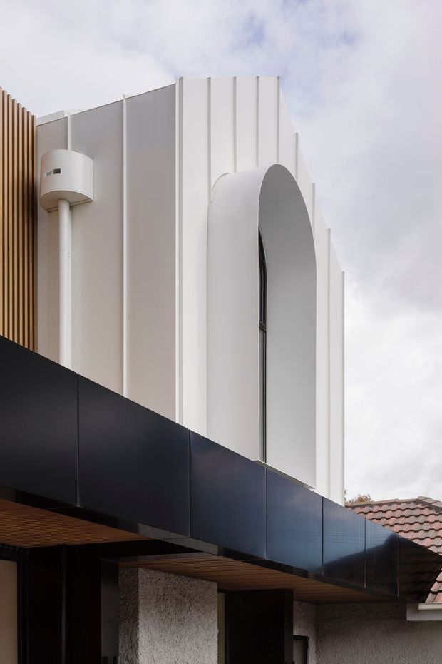 Exterior architectural view of a contemporary building facade, featuring a sculptural white upper volume with vertical ribbed detailing and a tall arched recessed opening. A projecting black metal-clad canopy or balcony spans the lower portion, with warm timber soffit lining and textured light-grey masonry walls below. A timber privacy screen and a small section of adjacent tiled roofing are visible.