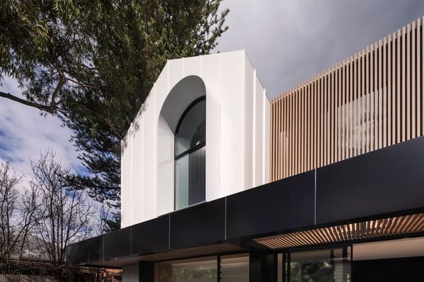 Exterior architectural view of a contemporary multi-level building with a crisp white facade, a tall arched window opening, dark charcoal cladding or screening, and a prominent upper-level timber-look vertical batten facade. The structure appears integrated into a leafy suburban setting beneath an overcast sky.