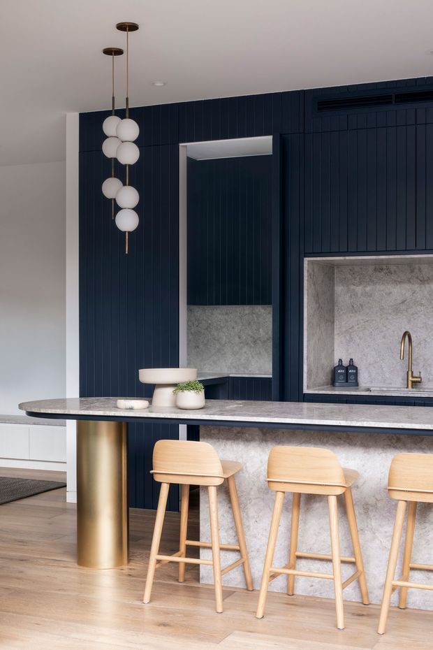 Contemporary open-plan kitchen featuring a deep navy vertically panelled wall and integrated cabinetry, a grey stone island with a cylindrical brass-toned support, pale timber bar stools, and a sculptural cluster pendant light. A stone-lined recessed sink area with brass tapware and a full-height mirror add depth and contrast. Light timber flooring and white cabinetry brighten the space.