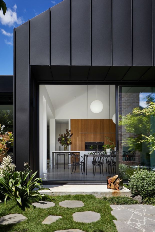 Contemporary house exterior with dark vertical metal cladding framing a large glazed opening into a bright dining and kitchen area. The interior features white walls, warm timber cabinetry, a dining table with black chairs, and spherical pendant lights. A landscaped garden with stepping stones and paved terrace sits in the foreground.